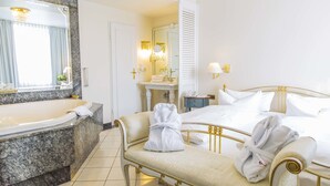 Habitación de hotel con baño de mármol, una cama grande con ropa blanca y una banca con bata.