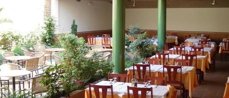 Un comedor interior con mesas y sillas, pilares verdes y plantas en macetas.