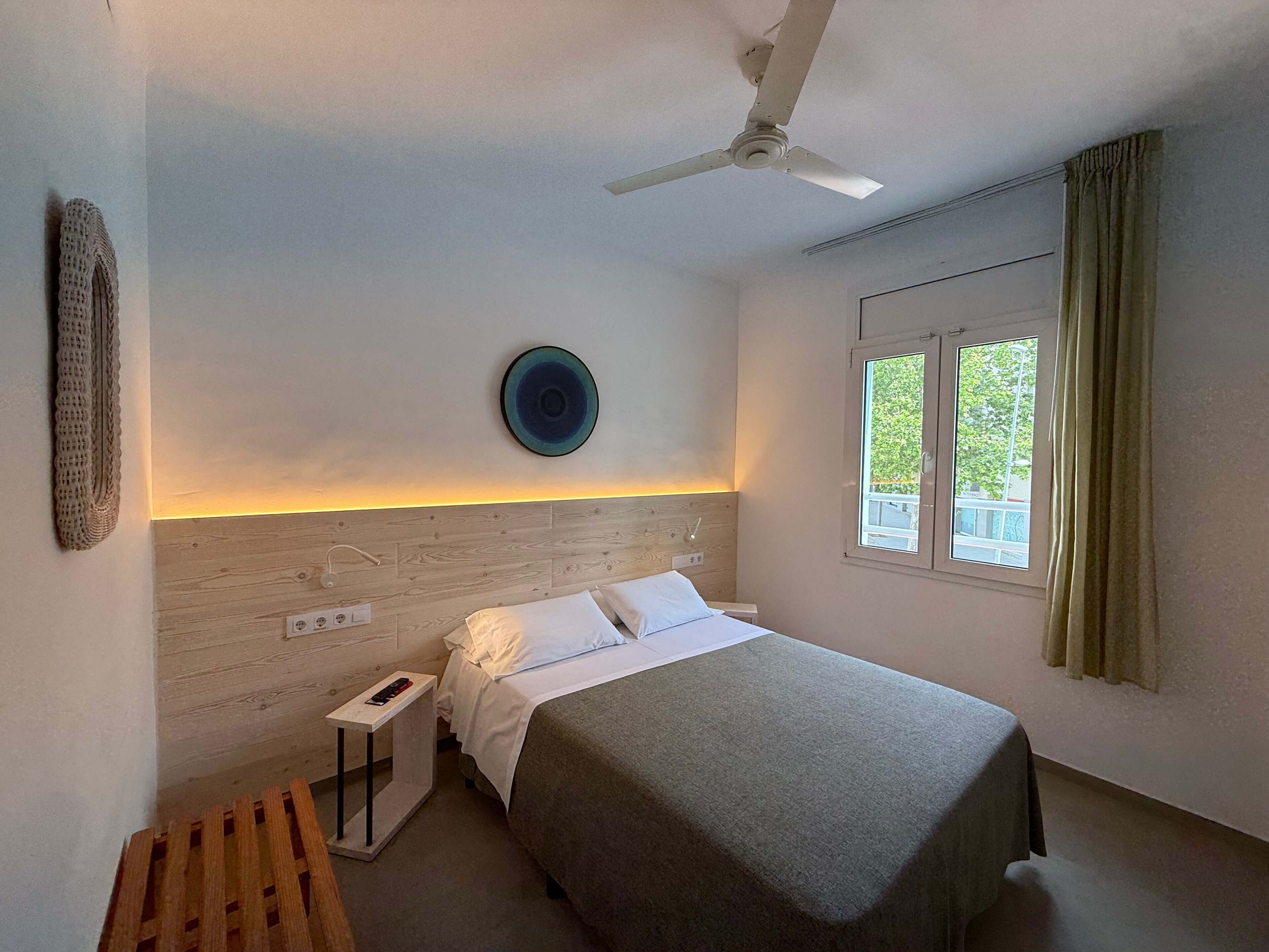 A bedroom with a bed, a ceiling fan, a bedside table, a nightstand, a window with curtains, and a wall-mounted plate.
