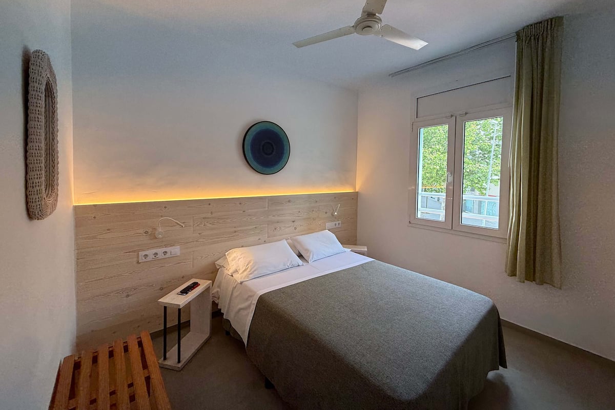 A bedroom with a bed, a ceiling fan, a bedside table, a nightstand, a window with curtains, and a wall-mounted plate.
