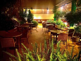 Un espacio al aire libre con mesas y sillas de mimbre, rodeado de macetas con plantas y helechos.