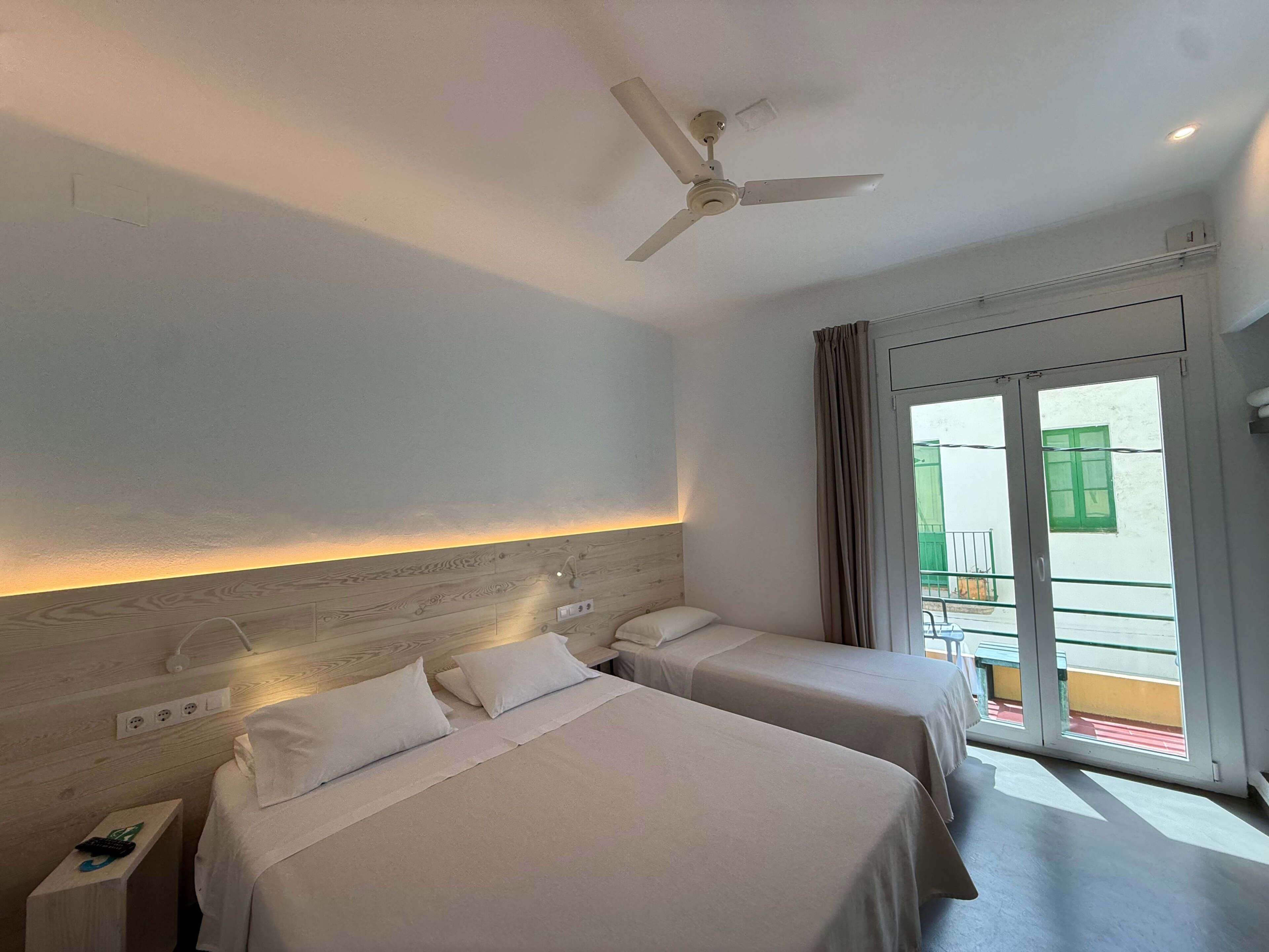 A hotel room with two beds, a ceiling fan, a window with a view of a balcony, and a door leading outside.