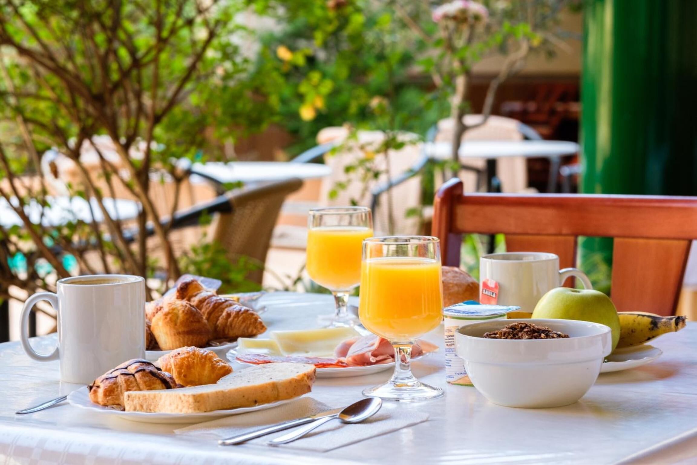 A breakfast table with croissants, toast, a bowl of cereal, a glass of orange juice, a coffee cup, and a banana.