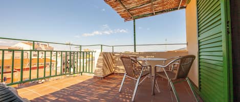 A terrace with a table and chairs, a green railing, and a view of buildings in the background.