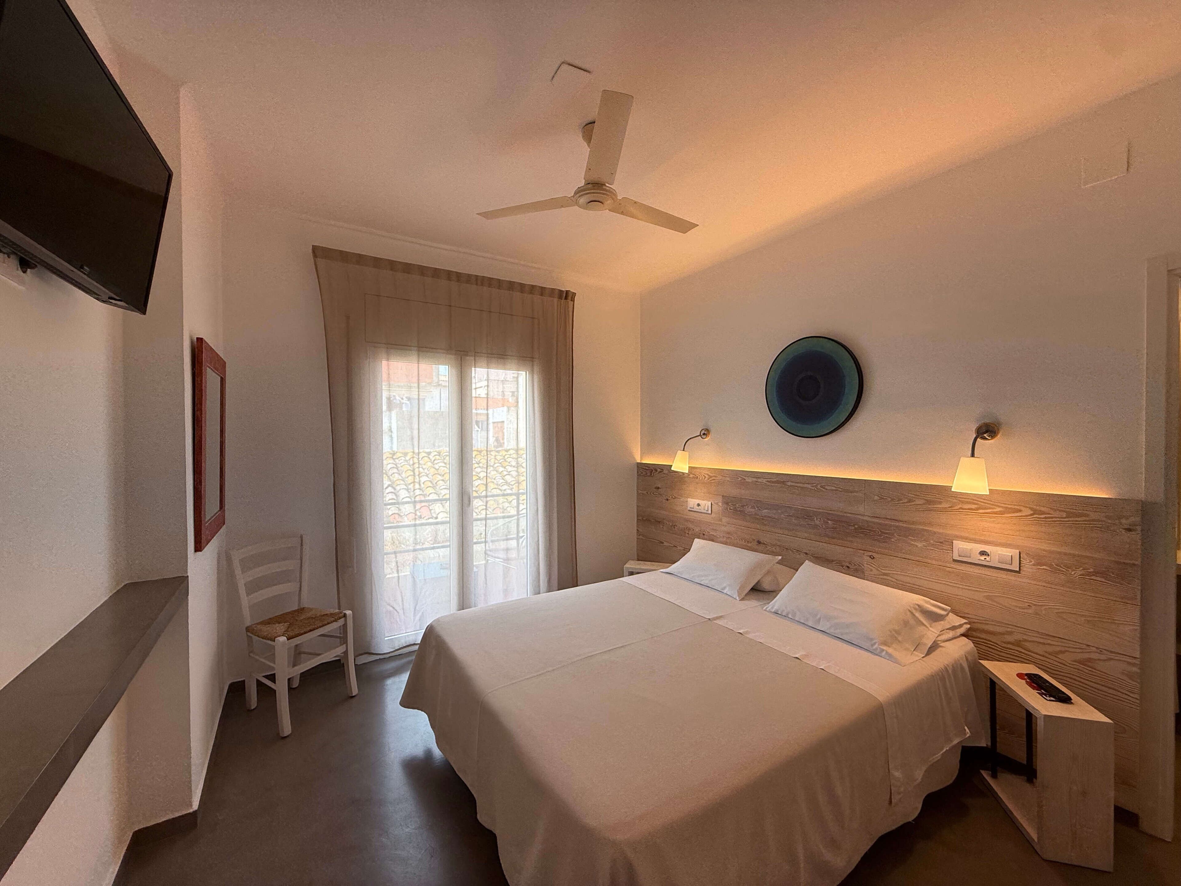 A hotel room with a bed, bedside tables, a ceiling fan, a TV, a chair, a window with curtains, and a wall-mounted lamp.