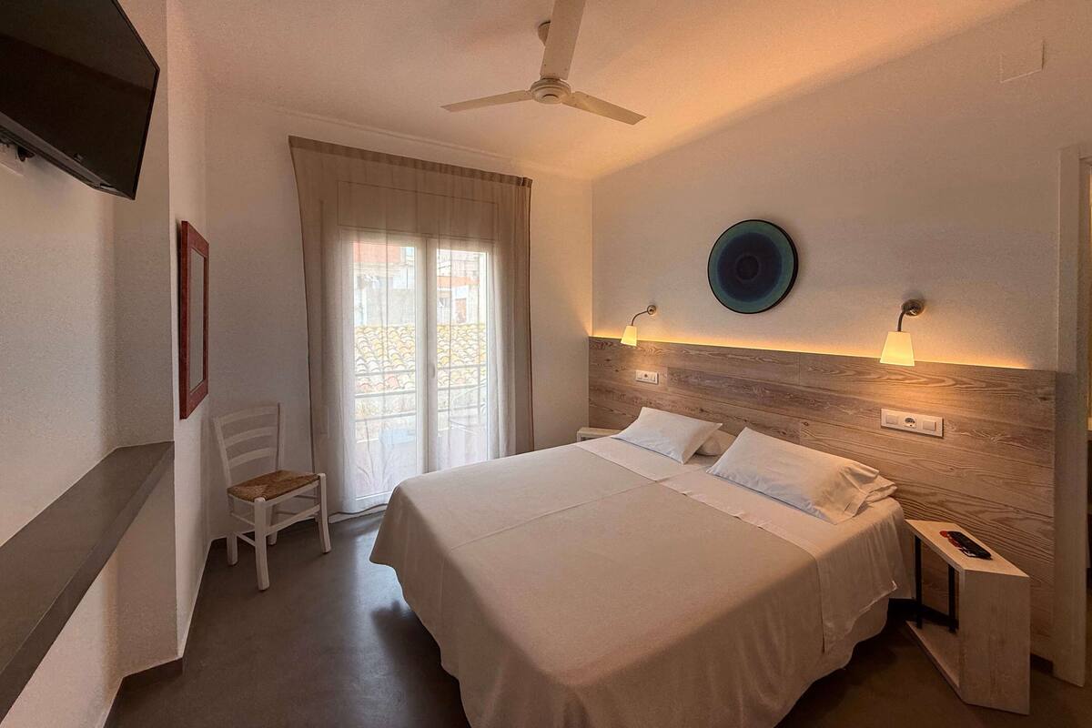 Une chambre d’hôtel comprenant un lit, des tables de chevet, un ventilateur de plafond, une télévision, une chaise, une fenêtre avec des rideaux et une lampe fixée au mur.