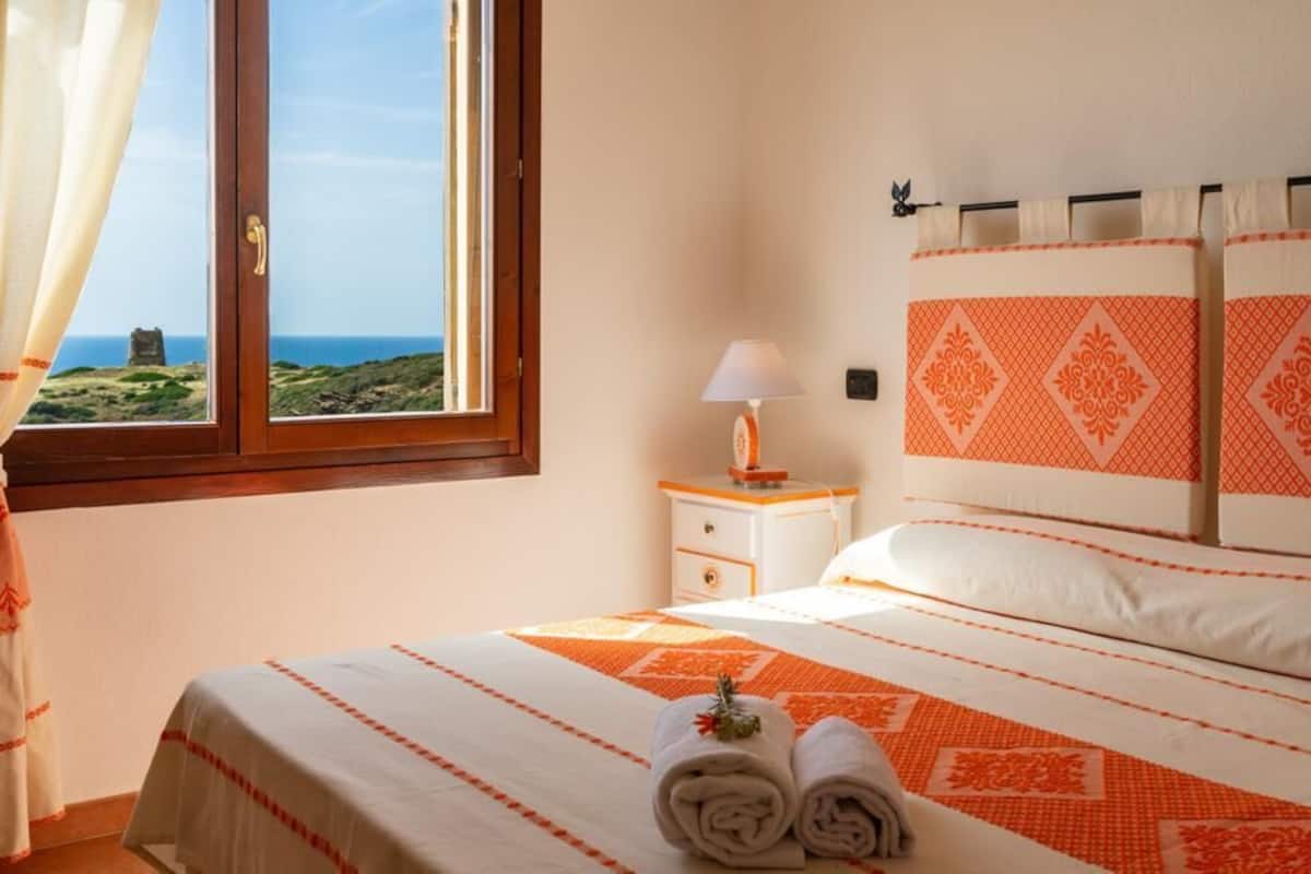 A bedroom with a bed, a nightstand, a lamp, and a view of the sea through a window.