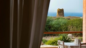 Vista attraverso una finestra con tende, che mostra una costruzione in pietra su una collina erbosa e un tavolo con sedie all'esterno.