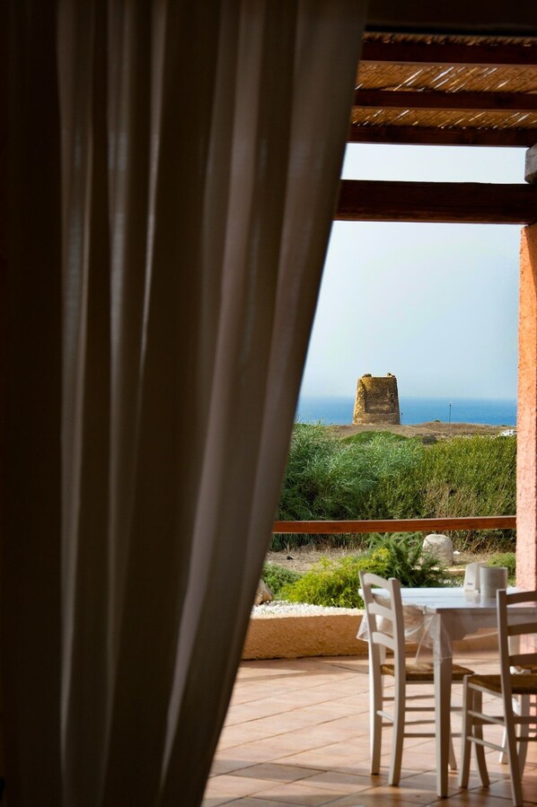 A view through a window with curtains, revealing a stone structure on a grassy hill and a table with chairs outside.