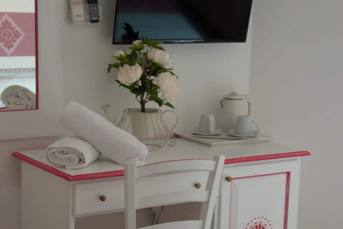 A white desk with a chair, a flat-screen TV mounted on the wall, a vase with flowers, and a small table with cups and a teapot.