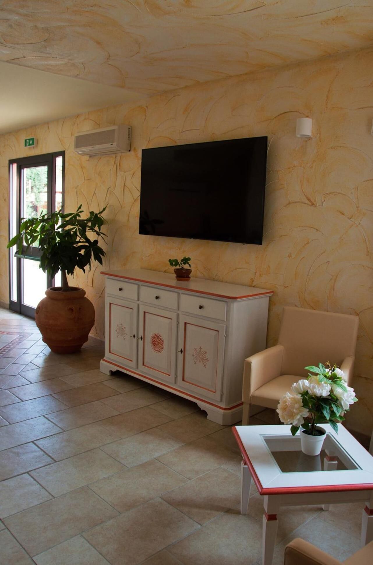 Una hall d'hotel con un'ampia televisione a schermo piatto montata a parete, un mobile bianco con ante decorate, una poltrona beige, un'ampia pianta in vaso e un tavolino con un vaso di fiori.