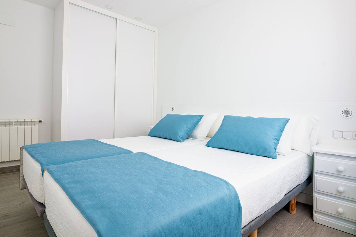 Una cama bien hecha con ropa de cama azul y blanca, un armario blanco y una mesita de noche con una lámpara.