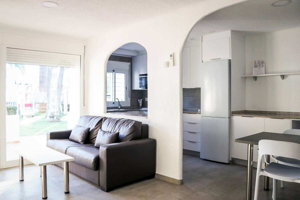 Una sala moderna con un sofá y una mesa de comedor, una cocina con armarios blancos, y un arco de entrada a otra habitación.