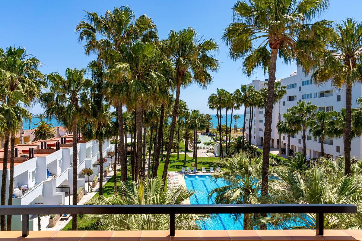 Un resort con piscina rodeada de palmeras, cielo azul despejado y vista al océano.