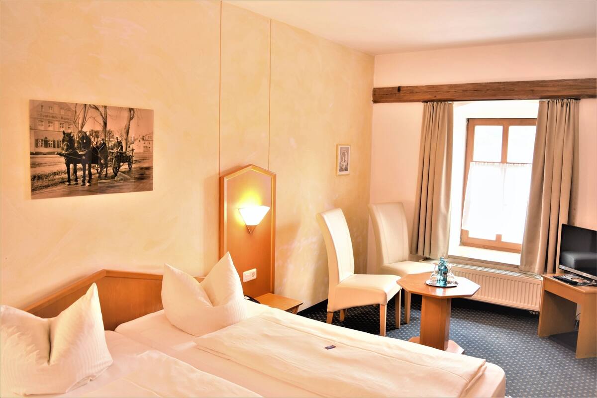 Una habitación de hotel con cama, una silla, escritorio, televisión y ventana con cortinas.