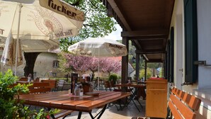 Un café en plein air avec des tables et des bancs en bois, abrité par de grands parasols arborant le logo « Tucher », entouré de plantes en fleurs et avec un bâtiment en pierre en arrière-plan.