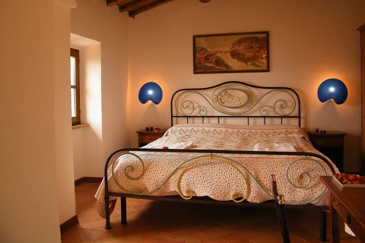 A bedroom with a metal bed frame, floral bedspread, two wall sconces, a painting above the bed, and a wooden ceiling.