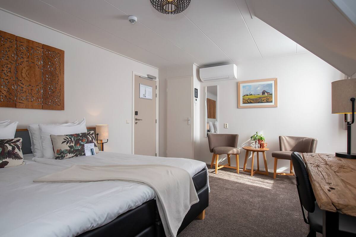 A hotel room with a bed, two chairs, a small table, a lamp, and a painting on the wall.