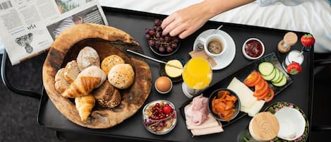 Bandeja de desayuno con cruasanes, panecillos, queso, jamón, huevos, uvas, zumo de naranja y diversas frutas y verduras.