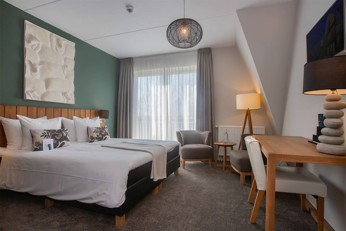 A hotel room with a large bed, a desk, a chair, a lamp, and a window with curtains.