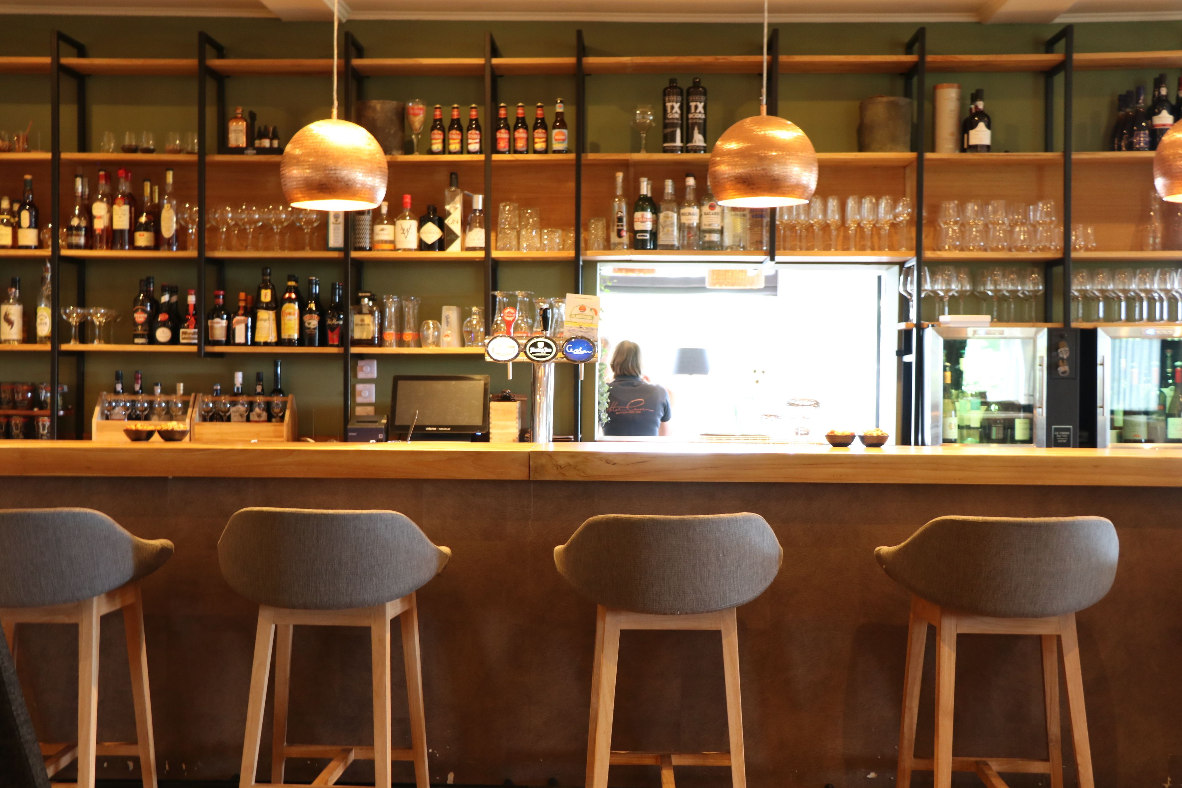 A bar area with wooden stools, shelves stocked with bottles and glasses, and pendant lighting.