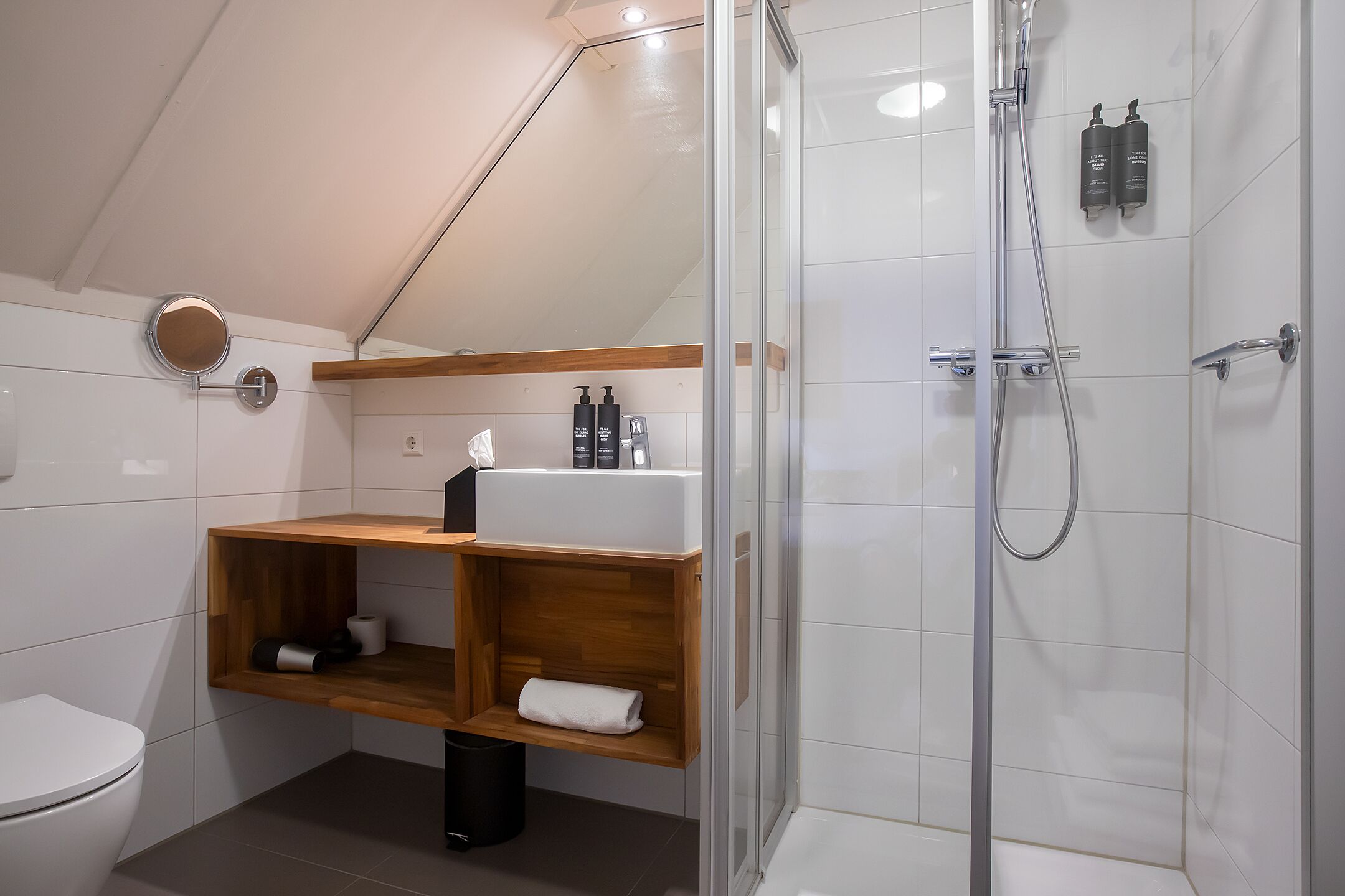 Une salle de bain moderne équipée d’une douche, de toilettes et d’étagères en bois où sont rangés les produits de toilette.