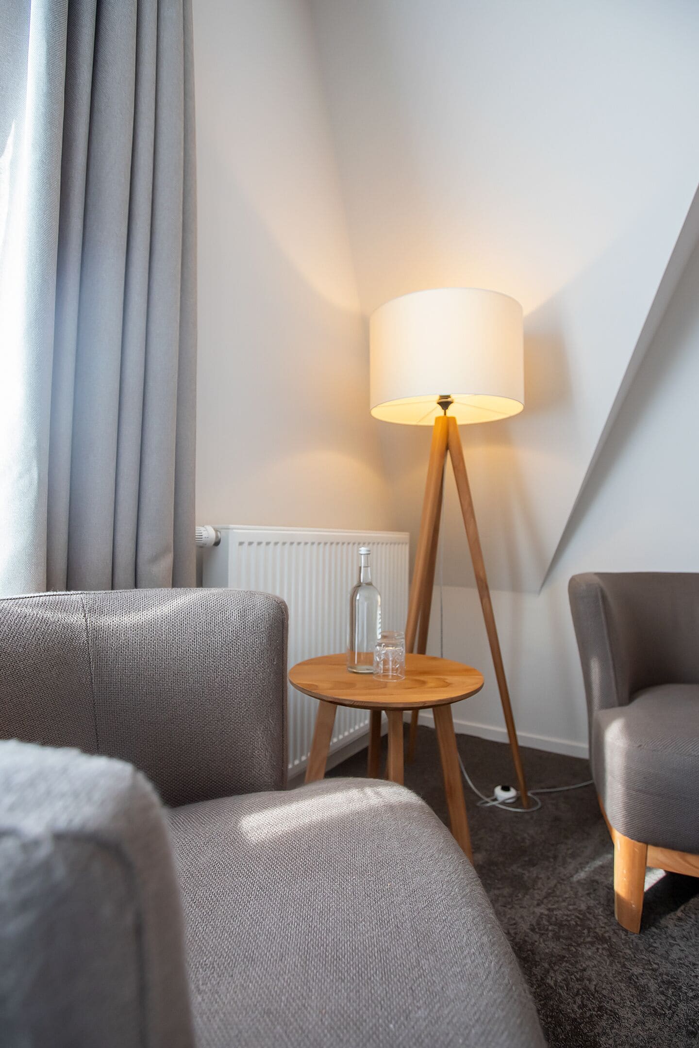 Une chambre d’hôtel moderne avec un canapé gris, un lampadaire en bois et une petite table ronde sur laquelle se trouvent une bouteille et un verre.