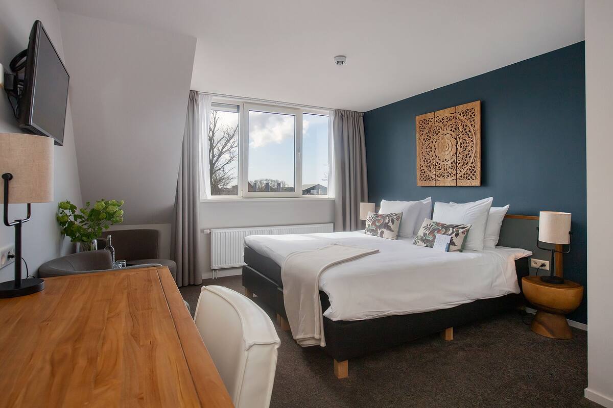 A hotel room with a bed, a desk, a chair, a TV, a window with curtains, and a decorative wall piece.
