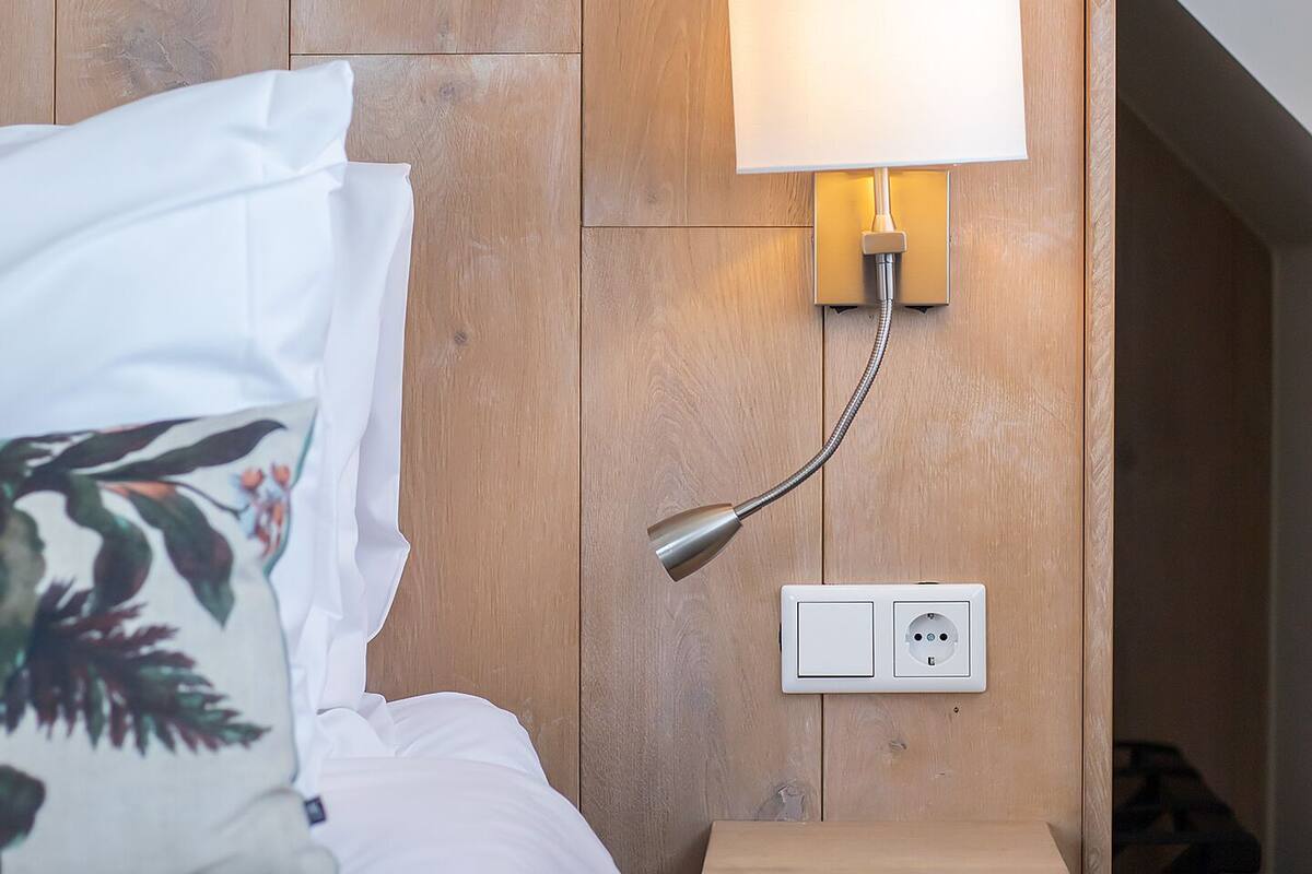 A bedroom with a wooden headboard, a bedside lamp, a power outlet, and a small wooden shelf.