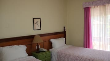 Habitación de hotel con dos camas, un cabecero de madera, una mesita de noche verde, una lámpara y una ventana con cortinas rosadas.