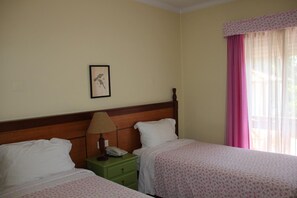 Habitación de hotel con dos camas, un cabecero de madera, una mesita de noche verde, una lámpara y una ventana con cortinas rosadas.