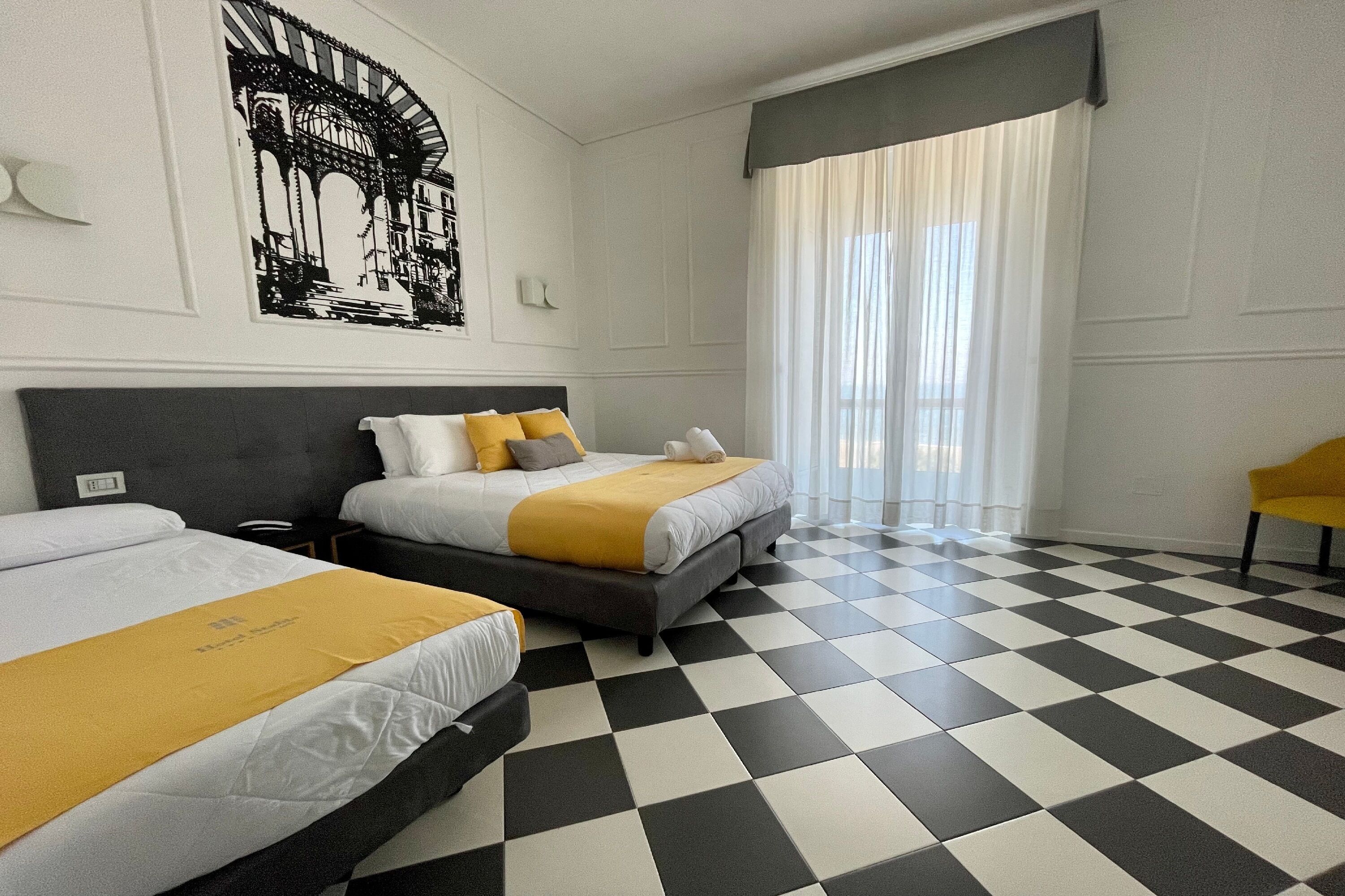 Een hotelkamer met twee bedden, een geruit vloertje, een groot raam met gordijnen en een gele stoel.