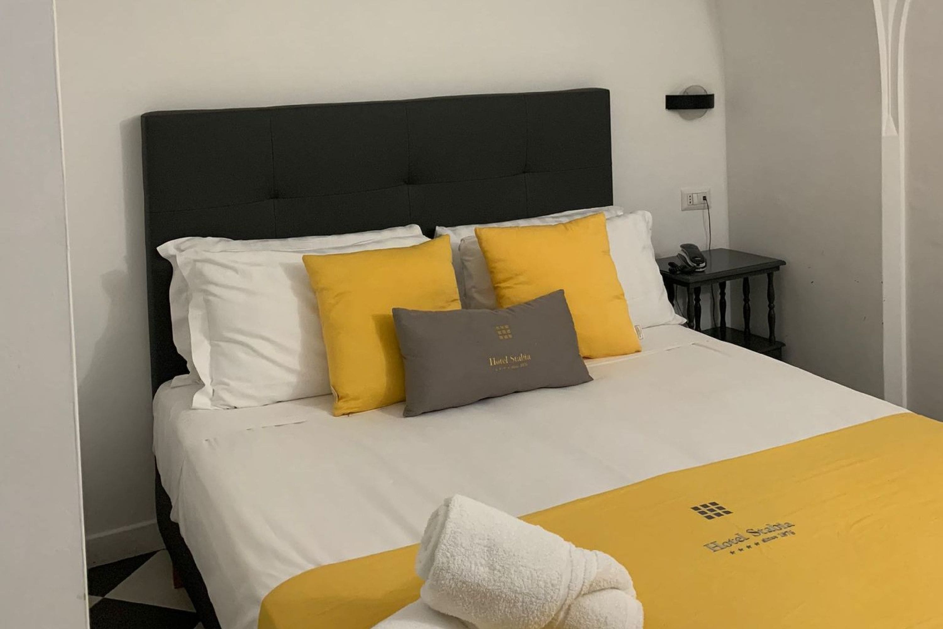 A neatly made bed with white and yellow bedding, a grey pillow, and a white towel on the bed.
