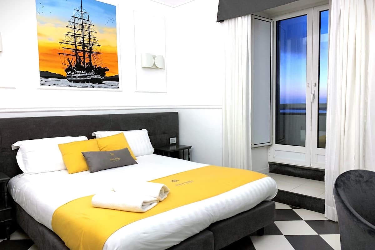 A hotel room with a large bed, a window with curtains, a painting of a ship on the wall, and a small table beside the bed.