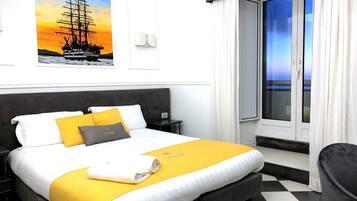 Una habitación de hotel con una cama grande, una ventana con cortinas, un cuadro de un barco en la pared y una mesita de noche.