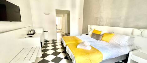 Une chambre d’hôtel avec un sol à carreaux, un lit avec une literie jaune et blanche, une table de chevet blanche et un téléviseur à écran plat fixé au mur.