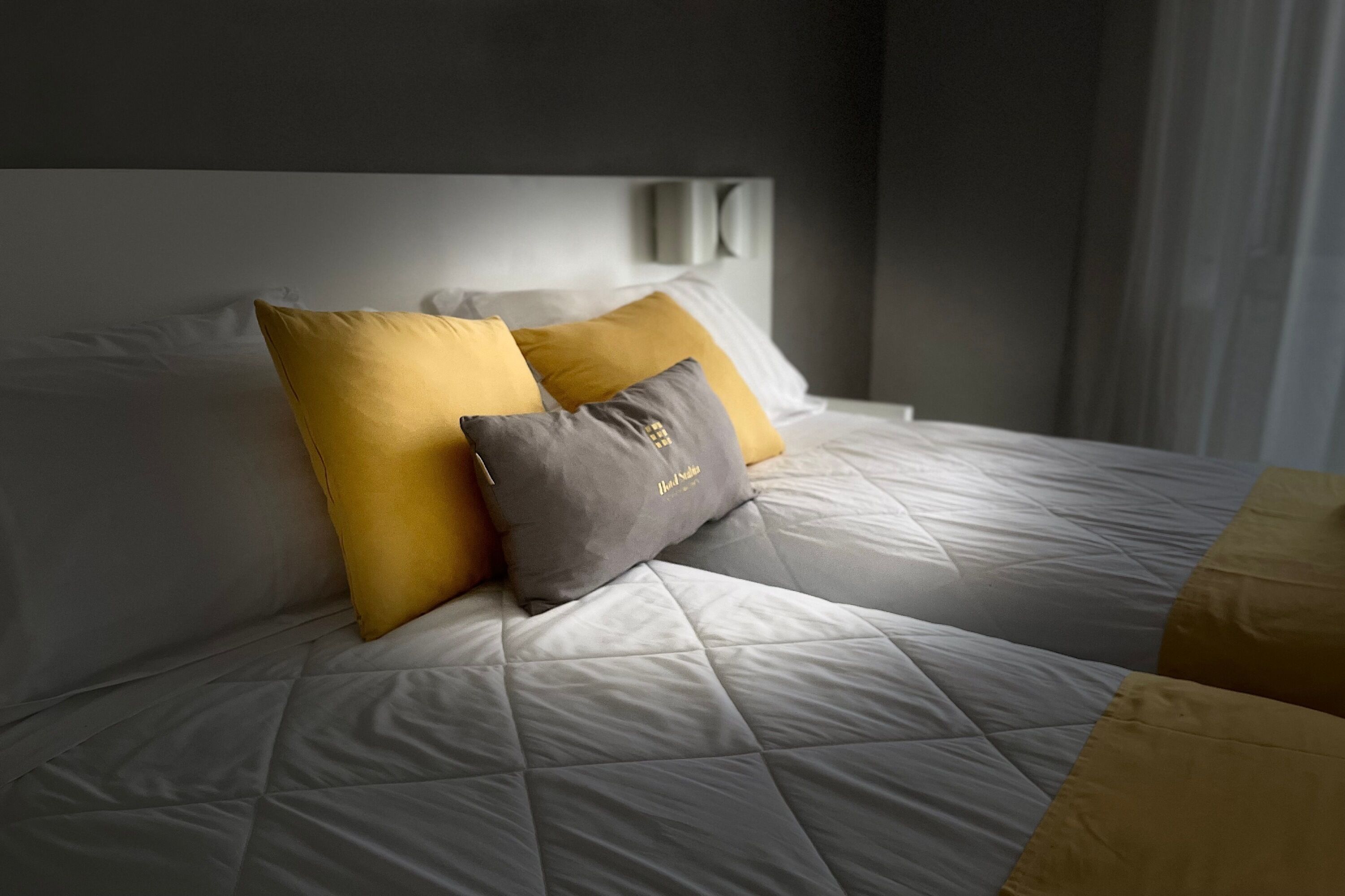 Una cama bien hecha con edredón blanco acolchado, dos almohadas en amarillo y gris, y un cabecero con una lámpara.
