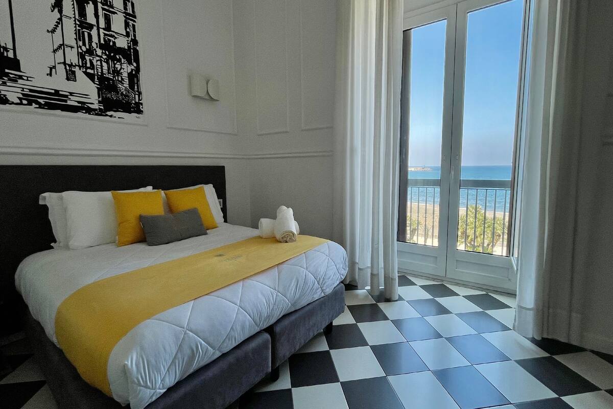 A hotel room with a checkered floor, a bed with a white and yellow bedspread, and a view of the sea through large windows.