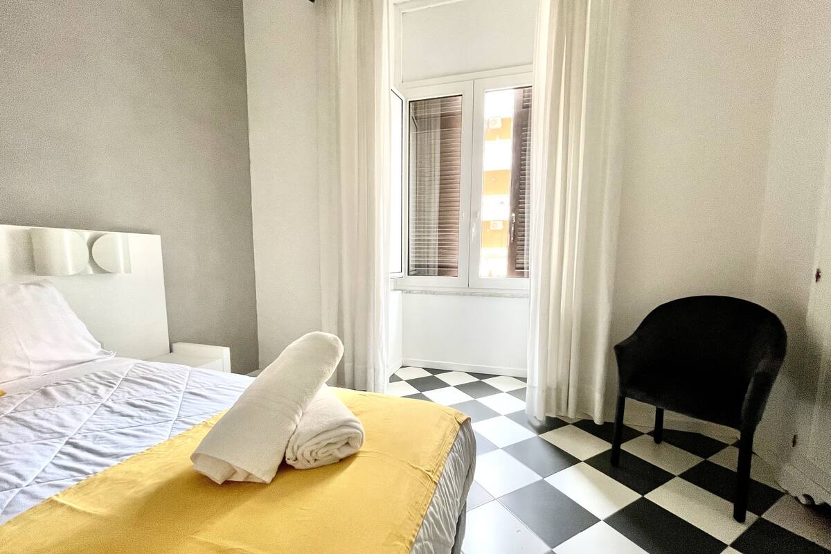 A hotel room with a checkered floor, a bed with a yellow blanket, white pillows, and a black chair.