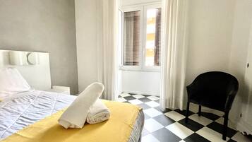 A hotel room with a checkered floor, a bed with a yellow blanket, white pillows, and a black chair.