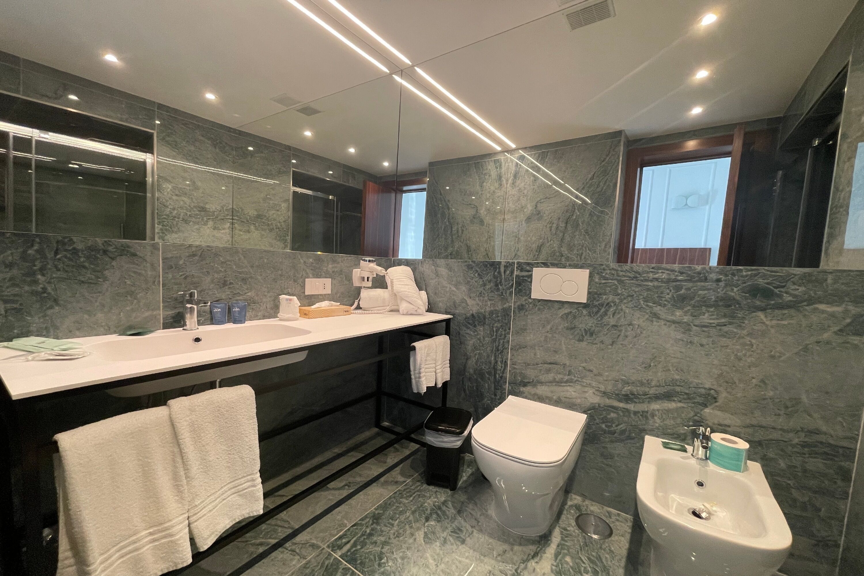 Ein modernes Badezimmer mit großem Spiegel, Waschbecken in Weiß, einem Bidet und einer Wand aus dunklem Marmor.