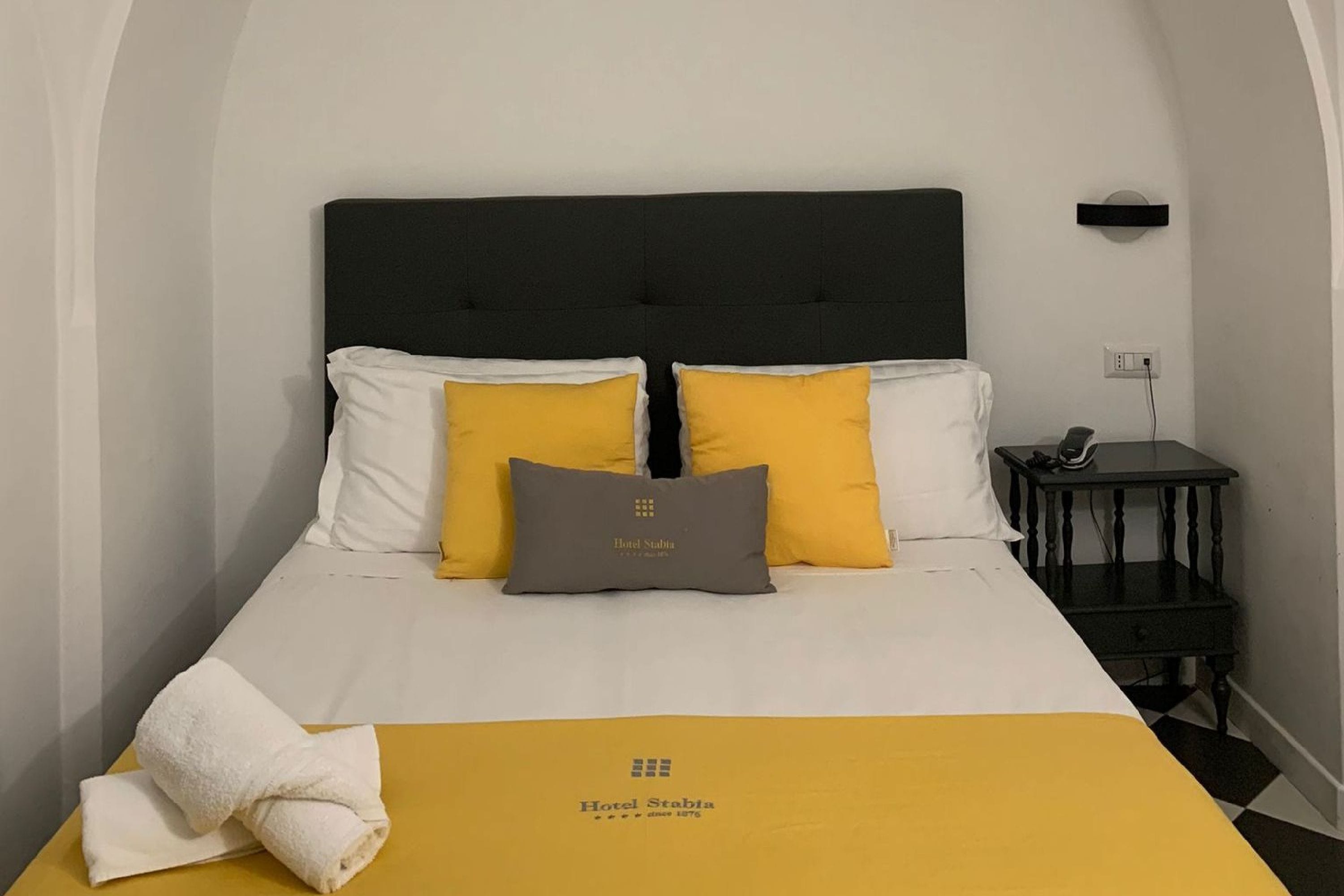 Een keurig opgemaakt bed met wit en geel beddengoed, een grijs kussen en een handdoek ernaast.