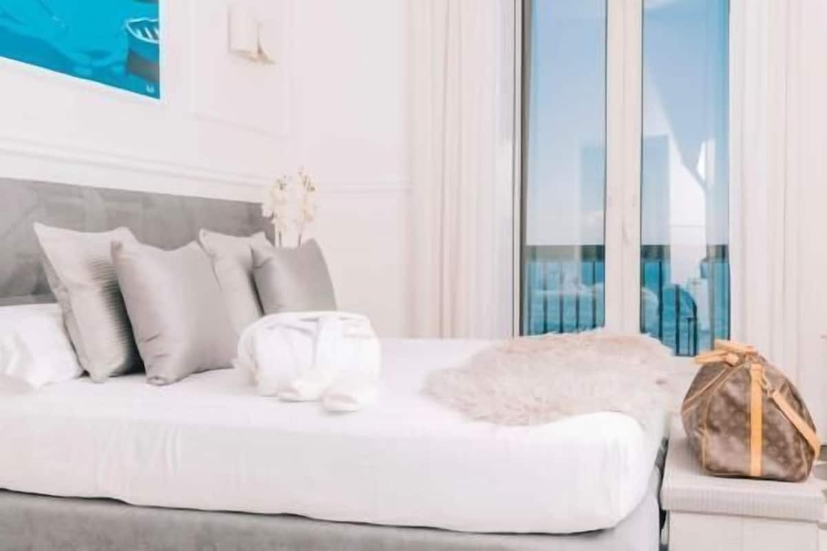 A bed with a grey headboard, white bedding, and a fluffy white pillow.