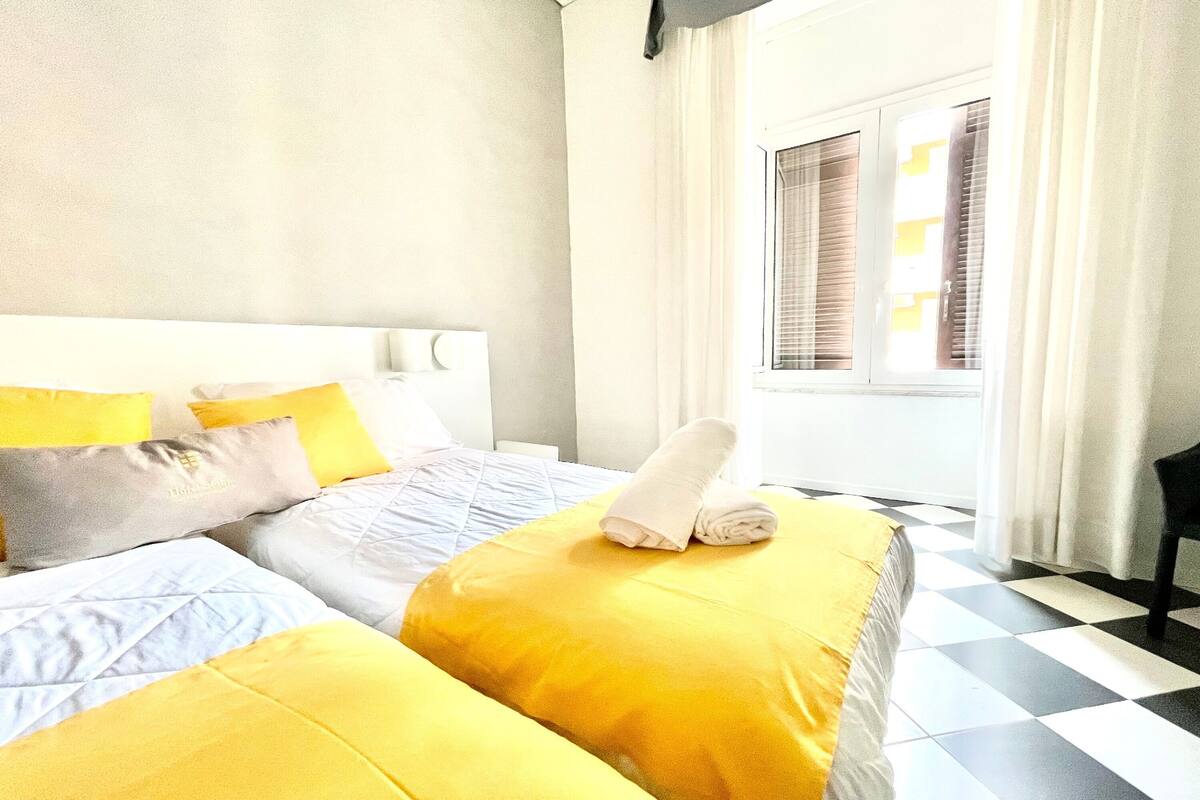 A hotel room with a checkered floor, a bed with yellow bedding, and a window with blinds.