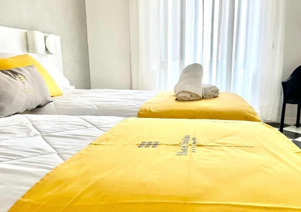 A hotel room with two beds, each with a yellow bedspread and a rolled towel on one bed.