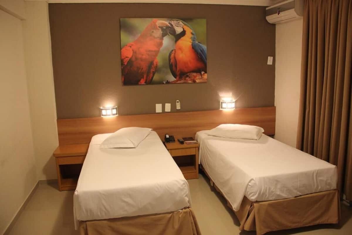 A hotel room with two single beds, a wooden headboard, a painting of parrots, bedside lamps, and a curtain.