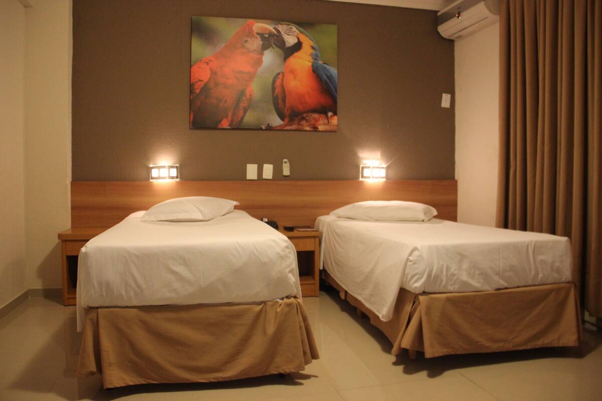 A hotel room with two single beds, a wooden headboard, a painting of parrots, and a nightstand with a lamp.