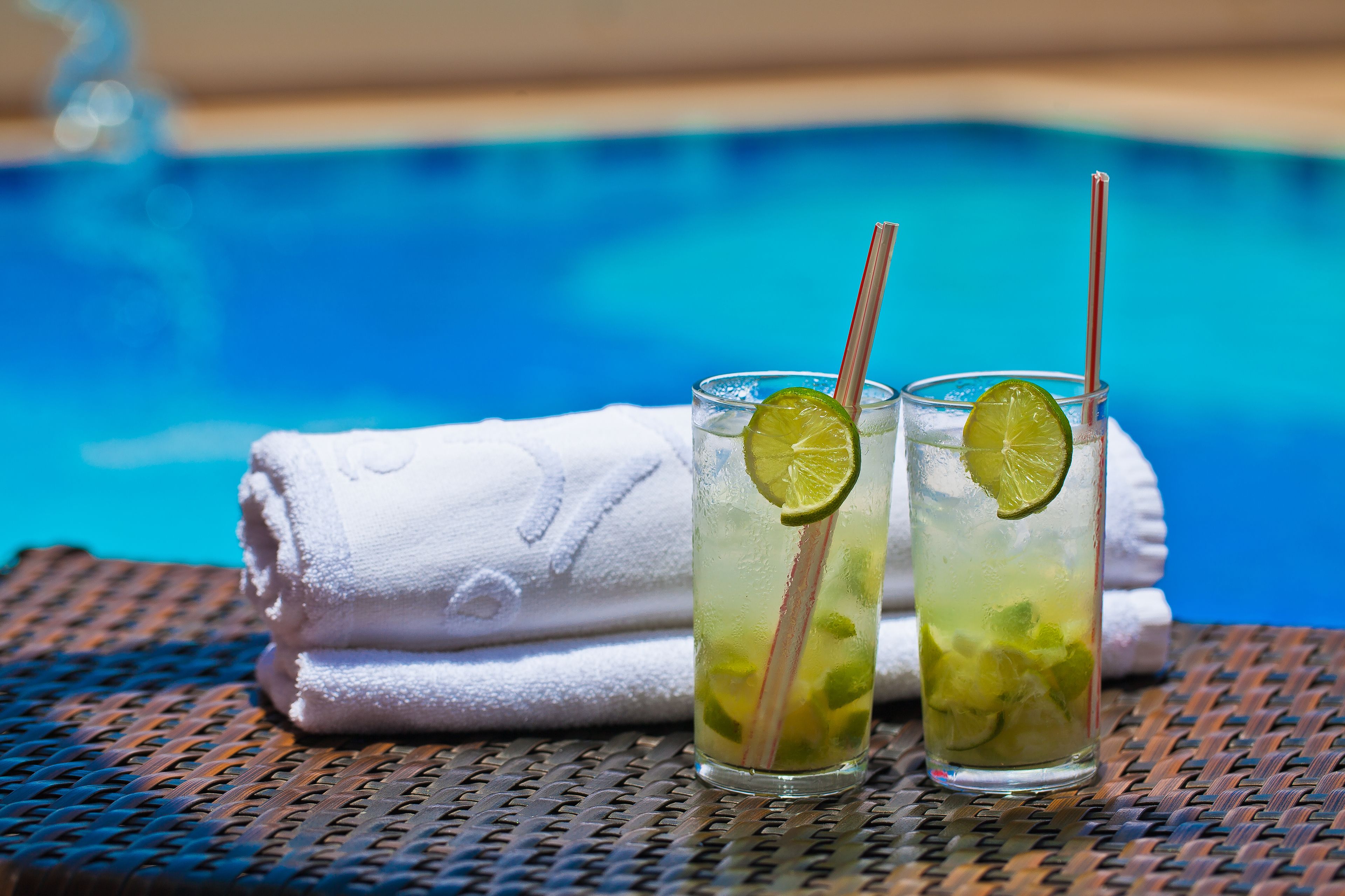 Two glasses of a lime-based beverage with straws, placed on a woven table beside a folded white towel by a pool.