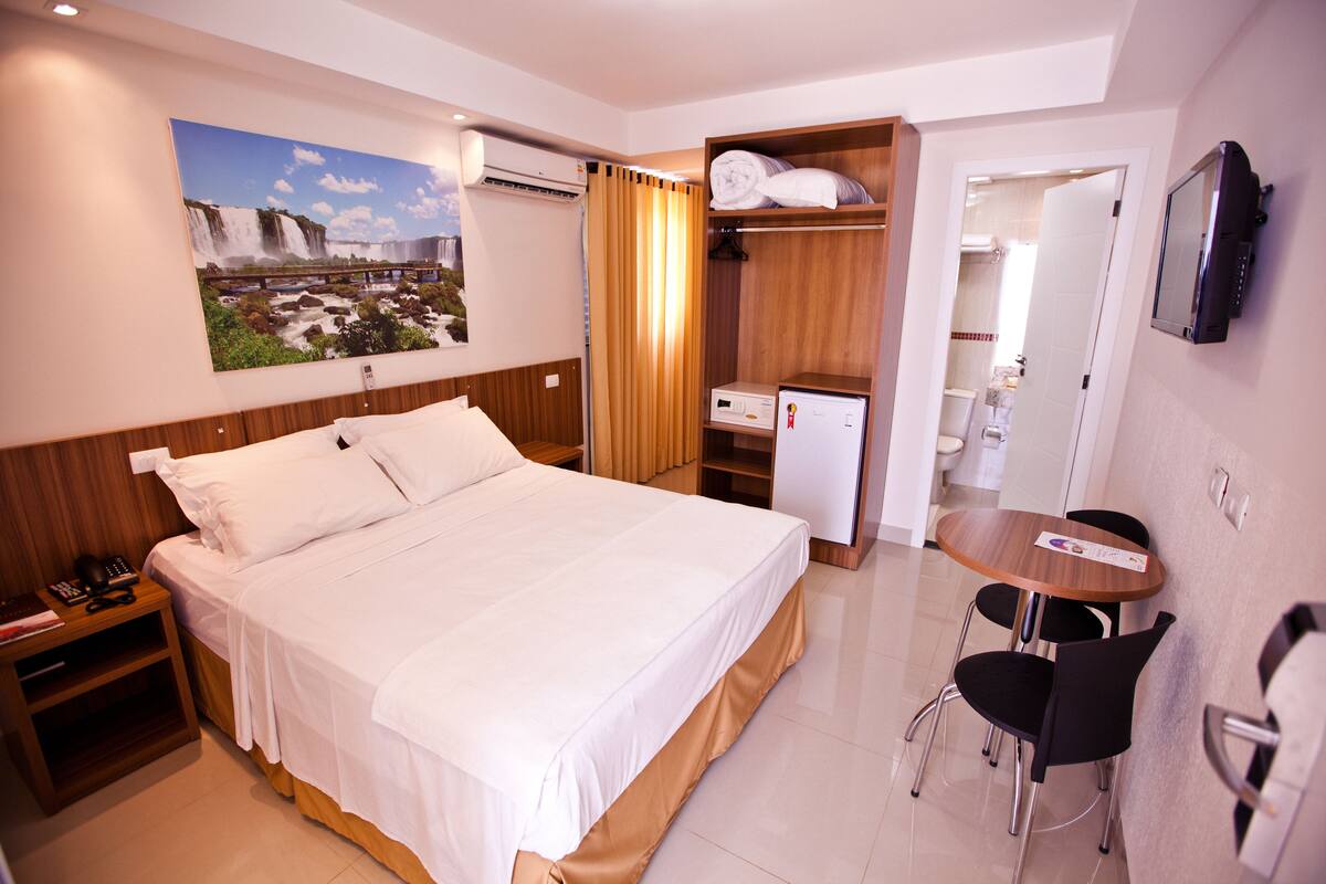 A hotel room with a bed, a desk with a chair, a television, a bathroom visible through an open door, and a painting on the wall.