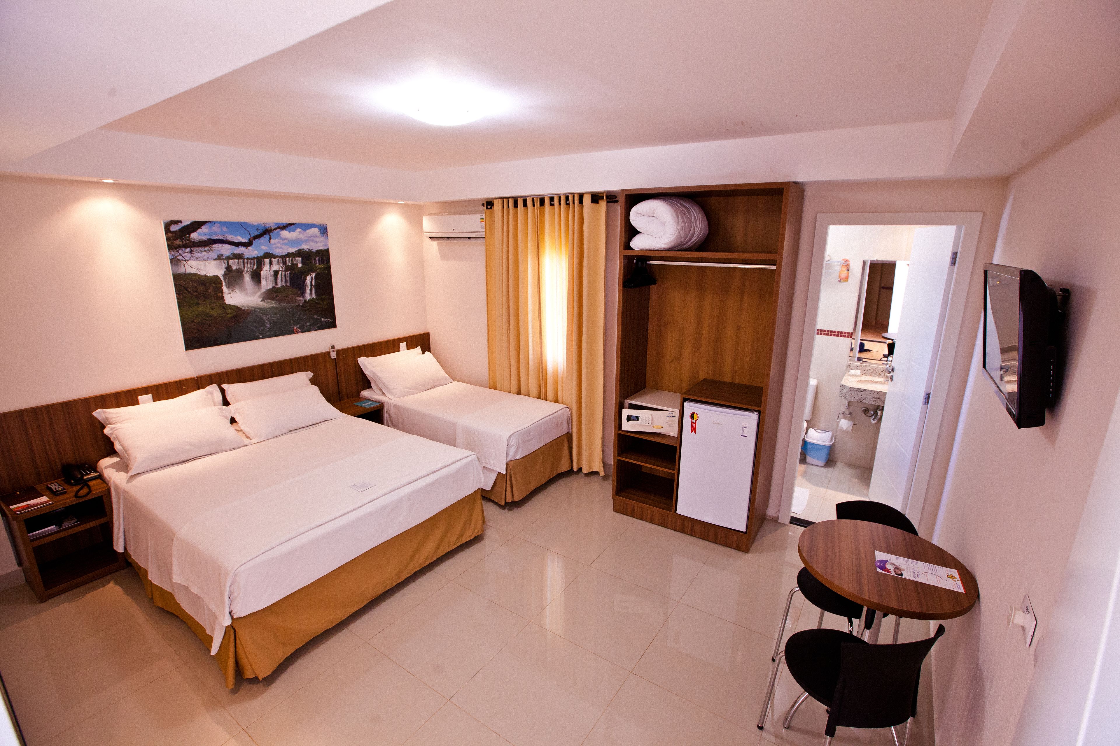 Habitación de hotel con dos camas, un escritorio, una silla, televisor, refrigerador y baño visible a través de una puerta abierta.